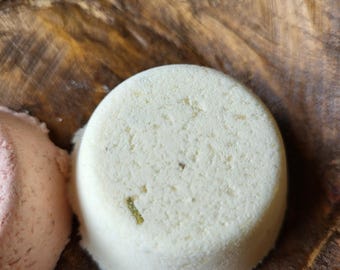 May include: A close-up shot of a white, round bath bomb with a textured surface. The bath bomb has small green flecks and sits on a wooden surface. A pink bath bomb is partially visible to the left.