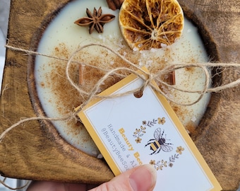 May include: A wooden bowl filled with a white candle with a cinnamon stick, dried orange slice, and star anise. The bowl is decorated with twine and a tag that reads "Beauty Bees Handmade & All Natural @BeautyBeesSoaps".