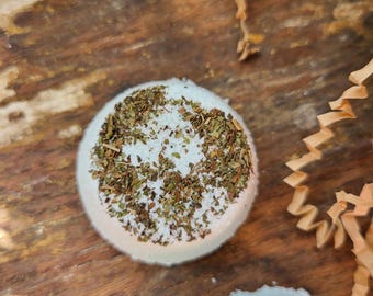 May include: Two round, white bath bombs with a light green hue, speckled with dried green herbs. One bath bomb is in focus, while the other is partially visible. The bath bombs rest on a rustic, wooden surface, with decorative paper accents.