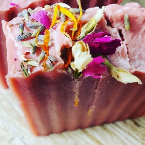 May include: A pink soap bar with dried flowers and herbs embedded in it. The soap is on a wooden surface.