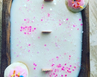 May include: A white candle with pink and iridescent glitter in a wooden bowl. The candle has three wooden wicks and is topped with two white cookies with pink frosting and colourful sprinkles.