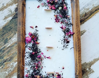 May include: A white candle with dried rose petals and tea leaves embedded in the wax. The candle is in a wooden rectangular tray.