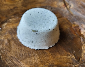 May include: A white, round bath bomb with flecks of herbs sits in a wooden bowl. The bath bomb has a slightly domed top and a cylindrical base. The wood grain of the bowl is visible, with varying shades of brown and a natural, organic texture.