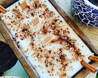 May include: A white candle with cinnamon, pecan pieces, and salt on top. The candle is in a wooden tray with a white flower on the front.