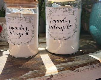 May include: Two glass jars of laundry detergent with wooden lids sit on a rustic wooden surface. The jars are labeled "All Natural Laundry Detergent" with a floral design. A small scoop rests inside each jar. The jars are filled with a white powder.