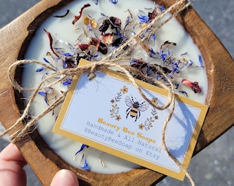May include: A round, white soap bar with dried flowers and herbs embedded in it. The soap is in a wooden, square-shaped dish. The soap is tied with twine and a tag that reads "Beauty Bee Soaps Handmade & All Natural @BeautyBeeSoap on Etsy".