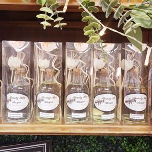 May include: A shelf display of eight clear glass bottles with white flower accents and wooden sticks. Each bottle has a label that reads "Beauty Bee Soaps".