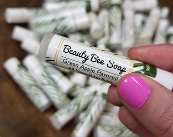May include: A close-up of a lip balm tube held by a person with pink nail polish. The tube is white with the text "Beauty Bee Soap" and "Green Apple Flavored All Natural Lip Balm". Other lip balms are in the background.