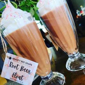 Puede incluir: Dos frascos de vidrio llenos de un líquido marrón cubierto con crema batida blanca y chispas rosas. Los frascos tienen forma de vasos de soda antiguos. Una etiqueta en uno de los frascos dice "Beauty Bee Soaps Handmade and All Natural Root Beer Float Hand Poured | 10 oz."