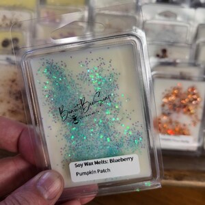 May include: A clear plastic container with a white soy wax melt inside. The wax melt is decorated with blue and purple glitter. The label on the container reads "Soy Wax Melts: Blueberry Pumpkin Patch".