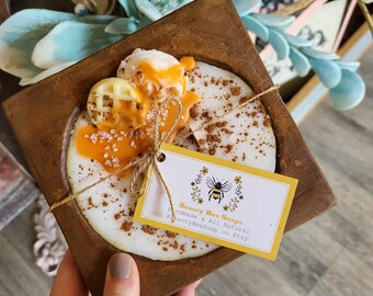 May include: A wooden tray with a white and yellow candle shaped like a waffle with a honey drizzle. The candle is on a white background with cinnamon sprinkled on top. A small tag is attached to the tray that reads "Beauty Bee Soaps Handmade & All Natural @BeautyBeeSoap on Etsy".