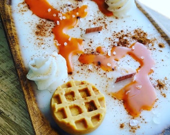 May include: A white candle with a brown wooden tray. The candle is decorated with two brown waffle-shaped candies, white frosting, and orange caramel sauce. The candle is sprinkled with brown cinnamon and white sugar.