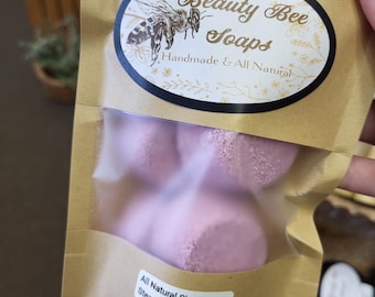 May include: A package of Beauty Bee Soaps handmade lavender shower steamers. The brown paper bag has a label with a bee illustration and the words "Beauty Bee Soaps Handmade & All Natural". The bag contains several pink, round shower steamers.