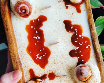 May include: A white candle in a wooden bowl with a brown, caramel-like sauce and white sprinkles. The candle has a cinnamon roll design.