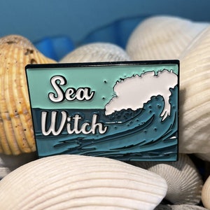May include: A teal blue enamel pin with a white wave and the text "Sea Witch" in white lettering.