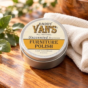 May include: A round silver metal tin of Daddy Van's All Natural Unscented Beeswax Furniture Polish. The label features the text Daddy Van's, Unscented Beeswax, Furniture Polish, Love Your Wood, and 6 ounces in black and gold.