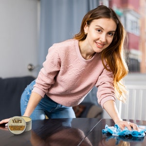 May include: A woman cleaning a dark wooden table with a blue and white cloth. A can of Daddy Van's Undead Furniture Polish is on the table. The can is brown and white with text.