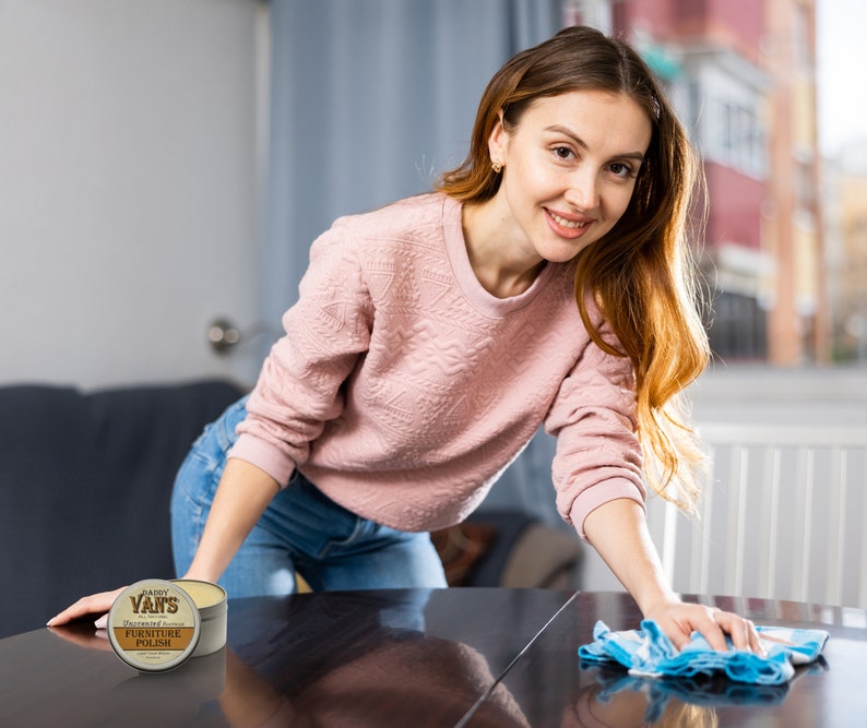 May include: A woman cleaning a dark wooden table with a blue and white cloth. A can of Daddy Van's Undead Furniture Polish is on the table. The can is brown and white with text.