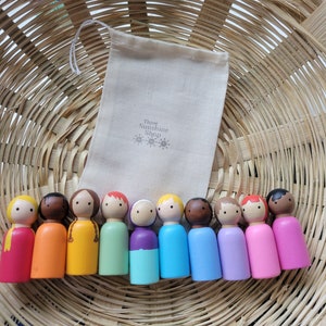 May include: A set of ten wooden peg dolls, each painted in a different colour of the rainbow. The dolls are arranged in a row in a wicker basket. The dolls are all wearing simple, painted faces. The dolls are packaged in a small white drawstring bag with the text "Three Sunshine Shop" printed on it.