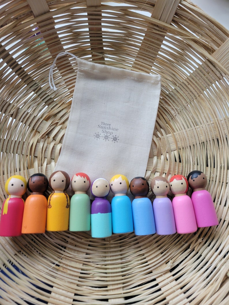 May include: A set of ten wooden peg dolls, each painted in a different colour of the rainbow. The dolls are arranged in a row in a wicker basket. The dolls are all wearing simple, painted faces. The dolls are packaged in a small white drawstring bag with the text "Three Sunshine Shop" printed on it.