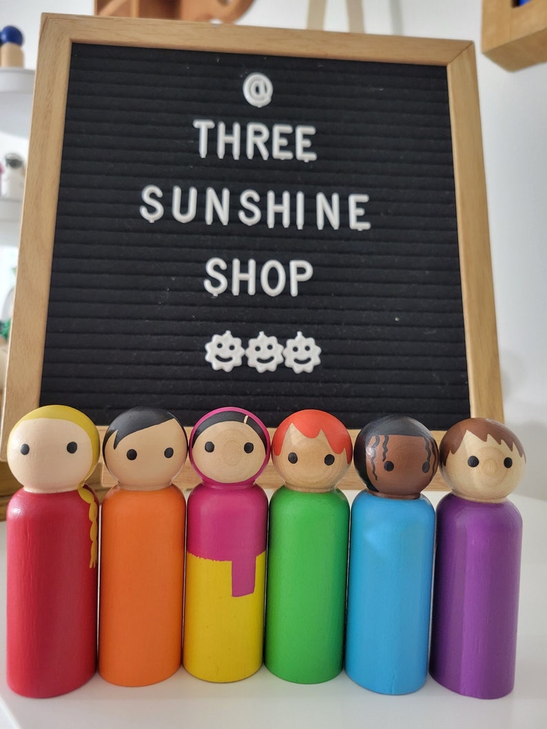 May include: Seven wooden peg dolls painted in different colors of the rainbow. The dolls are standing in a row on a white surface. The dolls have black eyes and mouths. The dolls are all the same size and shape. The dolls are a craft item.