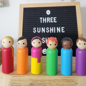 May include: Seven wooden peg dolls, each painted in a different color of the rainbow. The dolls are standing in a row on a wooden surface. The dolls have painted faces and hair.