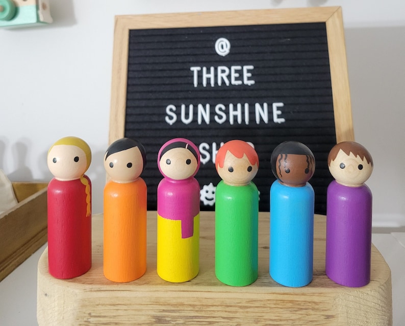 May include: Seven wooden peg dolls, each painted in a different color of the rainbow. The dolls are standing in a row on a wooden surface. The dolls have painted faces and hair.