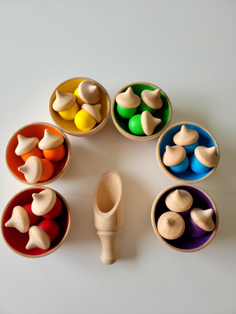 May include: A collection of wooden toys, including seven small bowls in various colors: yellow, green, orange, red, blue, and purple. Each bowl contains wooden acorn-shaped toys. A wooden scoop is also present.