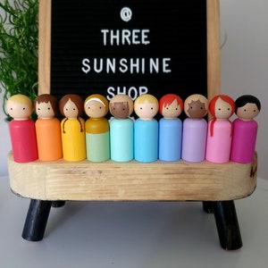 May include: A set of 12 wooden peg dolls painted in rainbow colours. The dolls are arranged in a row on a wooden stand with black legs. The dolls are all the same size and shape, and they have painted faces and hair.