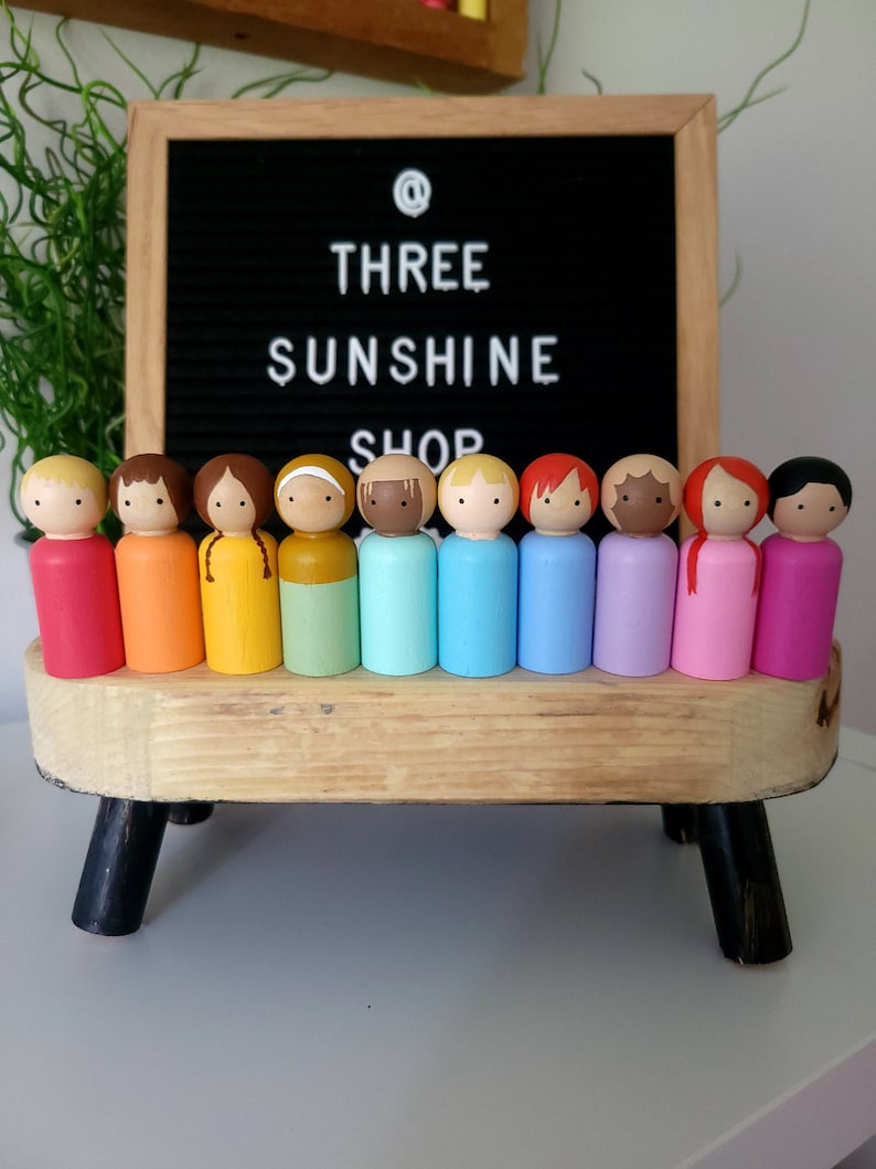May include: A set of 12 wooden peg dolls painted in rainbow colours. The dolls are arranged in a row on a wooden stand with black legs. The dolls are all the same size and shape, and they have painted faces and hair.