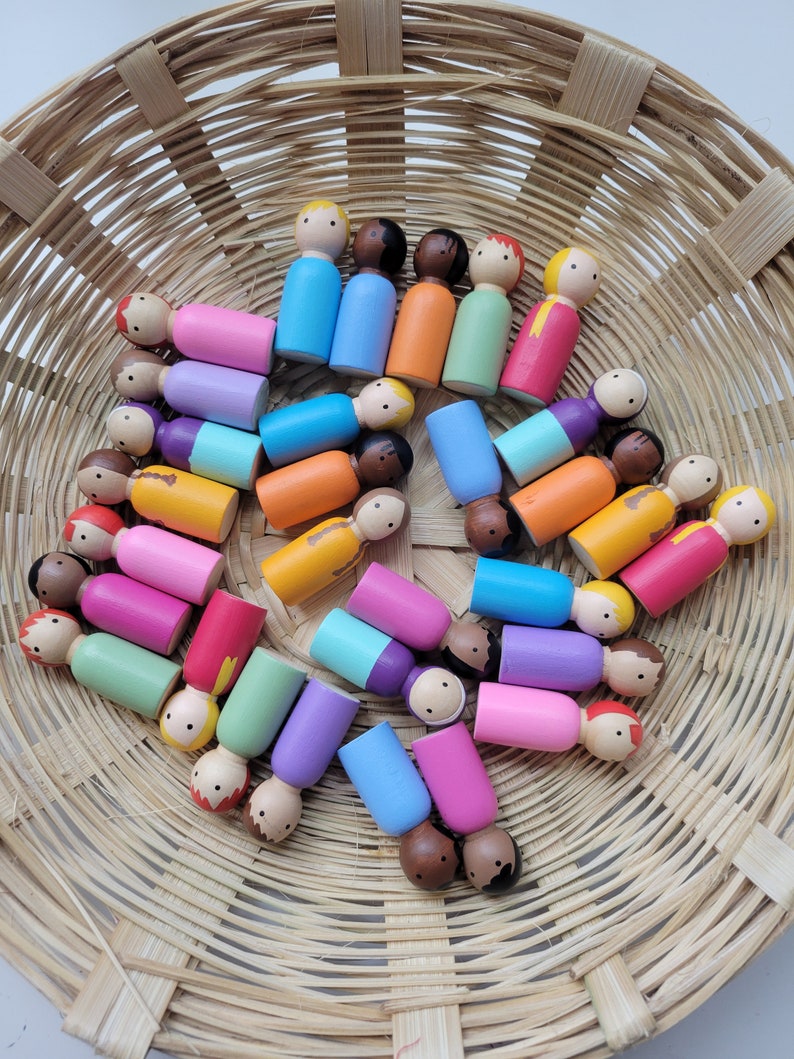 May include: A basket filled with colourful wooden peg dolls. The dolls are painted in various shades of pink, blue, green, orange, yellow, and brown. The dolls are arranged in a circular pattern.