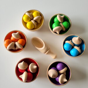 May include: A collection of colorful wooden acorn toys in small bowls. The bowls are yellow, green, orange, blue, red, and purple. A wooden scoop is in the center. The acorns have natural wood caps and painted bodies, ideal for educational play.
