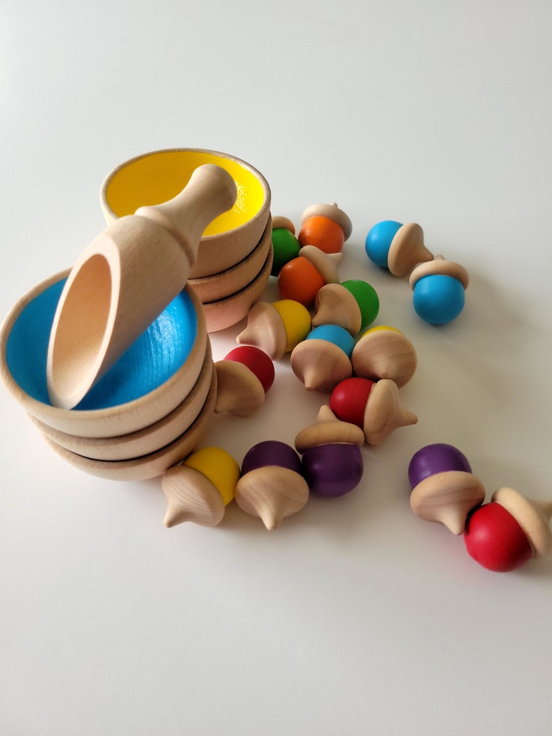 May include: Wooden toy set featuring small bowls with blue and yellow interiors, a wooden scoop, and colorful acorn-shaped pieces. The acorns have caps and painted tops in various colors, including red, purple, and blue. The set is designed for children.