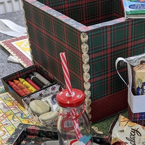 May include: A Christmas-themed gift box filled with various items, including a glass bottle with a red lid and a straw, a chocolate bar, a box of cookies, and a small box with a plaid design. The box is sitting on a table with a plaid tablecloth.