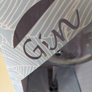 Könnte beinhalten: Ein klares Gin-Glas aus Glas mit einem silbernen Stiel, verpackt in einer blauen und weißen Schachtel mit einem Ausschnitt, auf dem "Gin" steht.