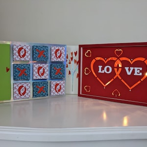 Op de afbeelding: Twee ingelijste kunstwerken met een Valentijnsdagthema. Het eerste stuk is een groene achtergrond met rode harten en een tic-tac-toe bord met rode X'en en blauwe O's. Het tweede stuk is een rode achtergrond met twee in elkaar verweven harten waarop in witte letters "LOVE" staat.