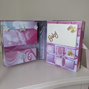 Könnte beinhalten: Ein rosa-weißes Baby-Scrapbook mit einer Holzfront. Das Scrapbook hat eine Schublade mit einem Elefantenmotiv und einen Bereich mit sechs kleinen Taschen. Das Scrapbook trägt das Wort "Baby" in goldenen Buchstaben und ein goldenes Fußabdruckdesign.