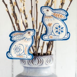 Two embroidered felt winter hare ornaments are hanging in branches of pussy willow in a grey vase. They are made of blue and white wool felt, with hand-embroidered details in a folk-art style.