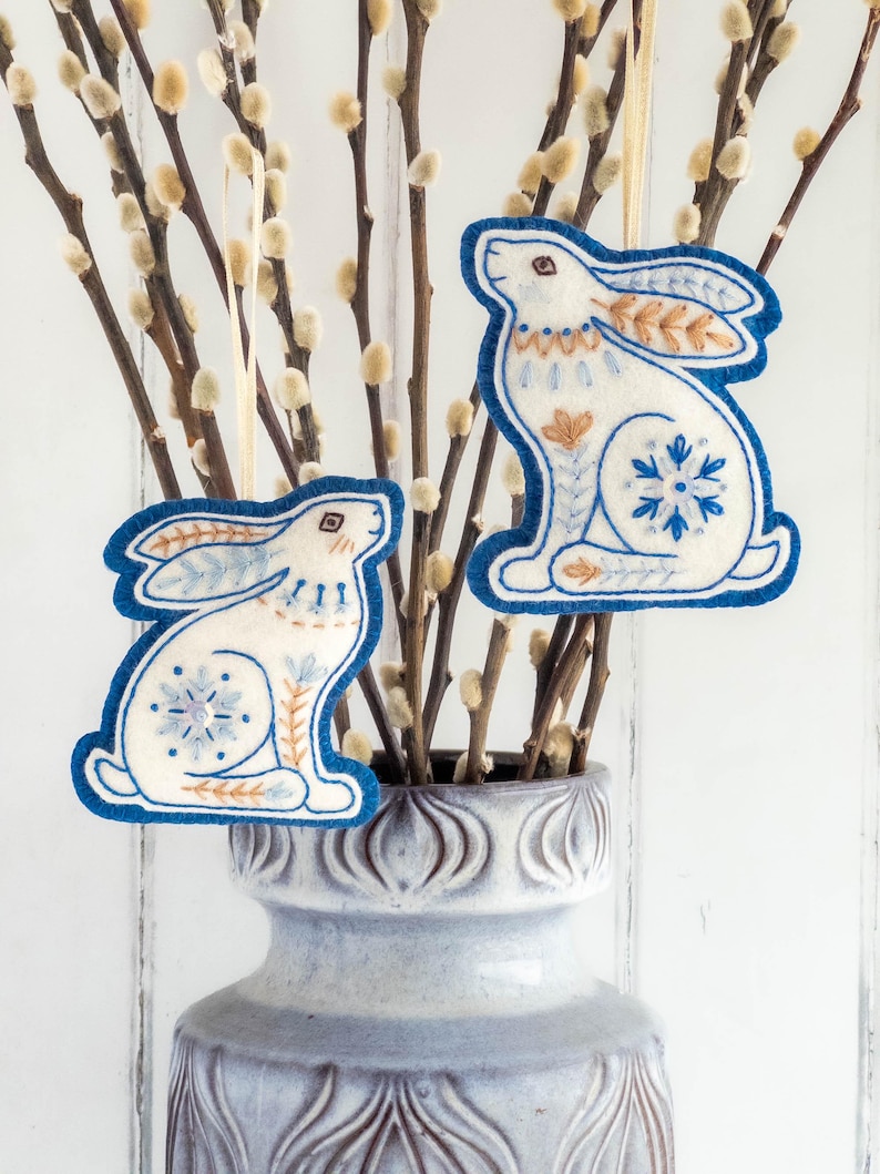 Two embroidered felt winter hare ornaments are hanging in branches of pussy willow in a grey vase. They are made of blue and white wool felt, with hand-embroidered details in a folk-art style.