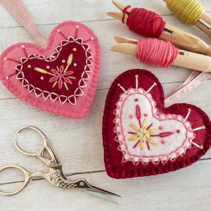 Two wool felt heart ornaments with pink and red embroidery. The hearts are decorated with gold thread and have a pale pink felt centre. The hearts are on a white wooden surface with pink and red thread spools and a pair of gold stork scissors.