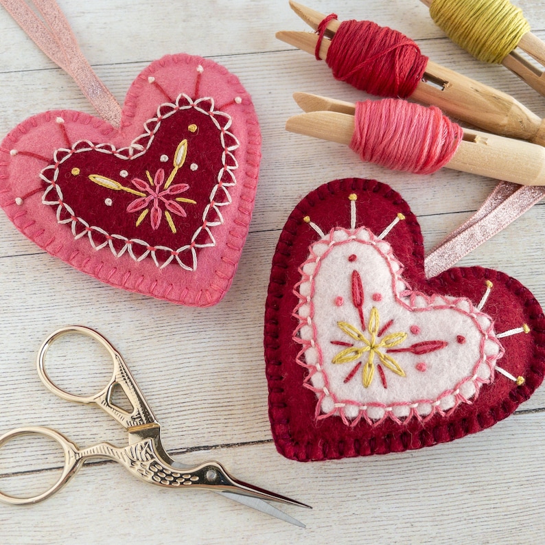 Two wool felt heart ornaments with pink and red embroidery. The hearts are decorated with gold thread and have a pale pink felt centre. The hearts are on a white wooden surface with pink and red thread spools and a pair of gold stork scissors.