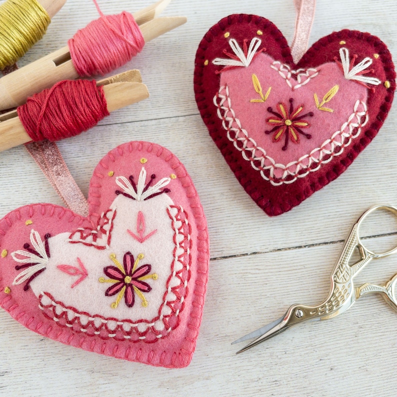 Two wool felt heart ornaments with pink and red embroidery are laying on a white-washed wooden surface. The embroidery pattern is in a folk-art style.