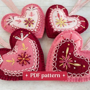 Four wool felt heart ornaments are laying on a white-washed wooden surface. The hearts have hand-embroidered details in red, white and pink and sparkly pink ribbon hangers. Overlaid text reads "PDF pattern".