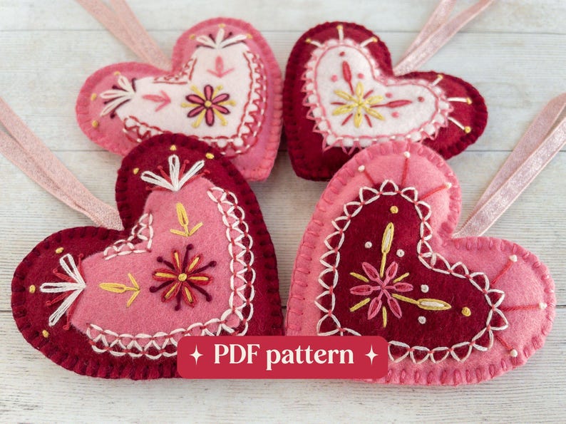 Four wool felt heart ornaments are laying on a white-washed wooden surface. The hearts have hand-embroidered details in red, white and pink and sparkly pink ribbon hangers. Overlaid text reads "PDF pattern".