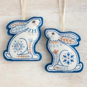 Two embroidered felt winter hare ornaments are arranged on a white-washed wooden surface. They are made of blue and white wool felt, with hand-embroidered details in a folk-art style, and they have gold ribbon hangers.