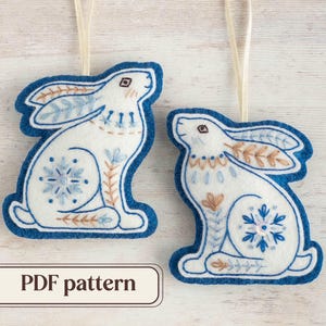May include: Two white felt ornaments shaped like rabbits with blue and brown embroidery. The ornaments have a snowflake design and are hanging from a white string.