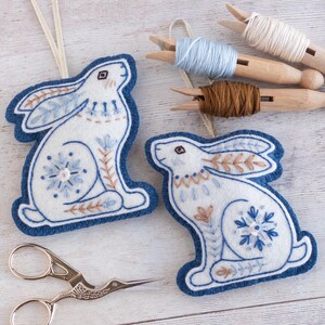 Two blue and felt winter hare ornaments with embroidered details are arranged on a white-washed wooden surface. They have hangers made of gold ribbon and snowflake motifs on their back legs. Embroidery thread and stork scissors are also in shot.