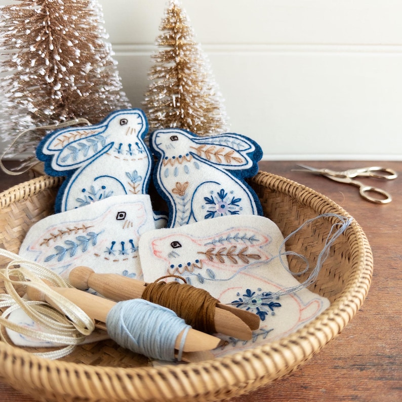 Four embroidered felt hare ornaments are in a wicker basket. Two are complete, the other two are a work in progress. Spools of embroidery thread and gold ribbon are also in the basket, and two Christmas tree ornaments are in the background.