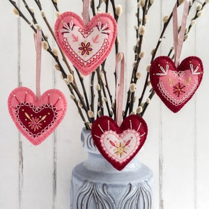 Four wool felt heart ornaments with embroidered details hang from a vase filled with pussy willow branches. The hearts are in shades of pink and red with white embroidery.
