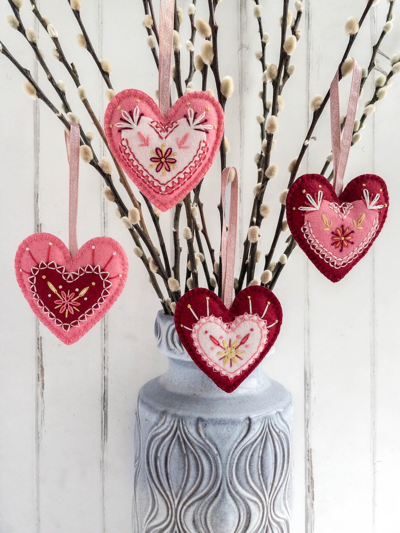 Four wool felt heart ornaments with embroidered details hang from a vase filled with pussy willow branches. The hearts are in shades of pink and red with white embroidery.